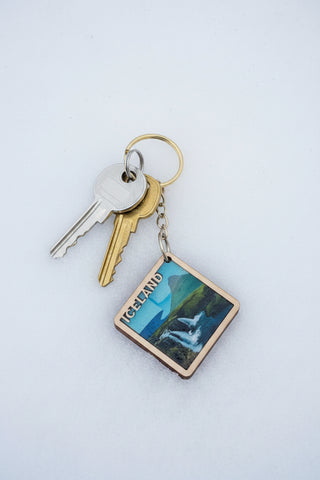 A square  keychain featuring a scenic landscape of Iceland with a waterfall and mountains, attached to a metal key ring and displayed on a brown cardboard backing card labeled 'Nordic Keychains'.