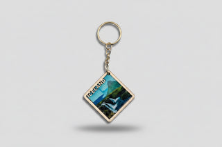 A square  keychain featuring a scenic landscape of Iceland with a waterfall and mountains, attached to a metal key ring and displayed on a brown cardboard backing card labeled 'Nordic Keychains'.