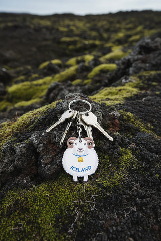 A sheep-shaped souvenir keychain from Nordic Souvenirs, featuring a white sheep with brown horns and a blue 'ICELAND' collar, attached to a gold key ring on a brown cardboard backing card.