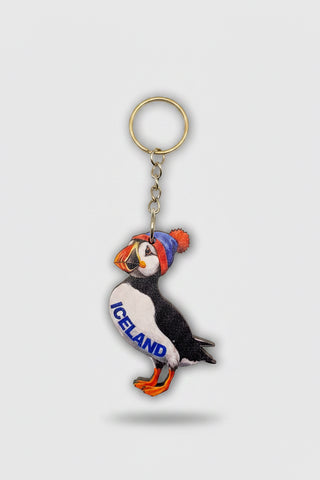A puffin-shaped souvenir keychain from Nordic Souvenirs, attached to a brown cardboard hanger card. The puffin is black and white with orange and blue accents, wearing a blue hat and standing on orange feet, with the word 'ICELAND' embroidered on its side.