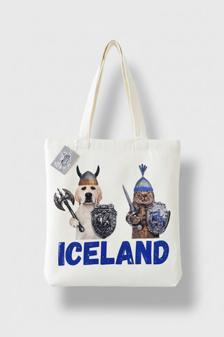 A white canvas tote bag with two cream-colored shoulder straps, featuring a graphic print of a dog dressed as a Viking on the left and a cat dressed as a warrior on the right, with the word 'ICELAND' printed in bold blue letters at the bottom.