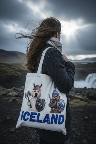 A white canvas tote bag with two cream-colored shoulder straps, featuring a graphic print of a dog dressed as a Viking on the left and a cat dressed as a warrior on the right, with the word 'ICELAND' printed in bold blue letters at the bottom.
