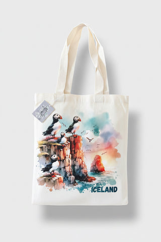 A white canvas tote bag featuring a colorful watercolor illustration of puffins on cliffs with the word 'ICELAND' printed at the bottom, shown against a light gray background.