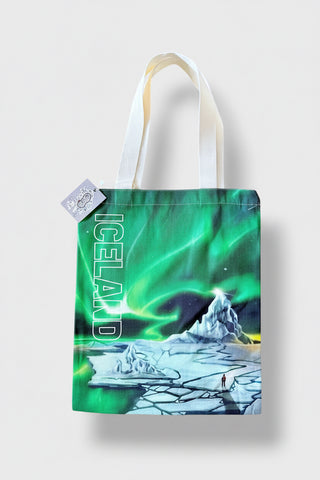 A canvas tote bag featuring a vibrant green aurora borealis print over an icy landscape with the word 'ICELAND' printed vertically in white. The bag has white fabric handles and a small brand tag on the side, set against a light gray background.