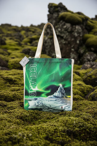 A canvas tote bag featuring a vibrant green aurora borealis print over an icy landscape with the word 'ICELAND' printed vertically in white. The bag has white fabric handles and a small brand tag on the side, set against a light gray background.