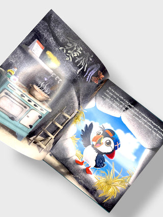 An open illustrated book or magazine spread photographed on a light gray background. The pages show a colorful cartoon puffin in a bright blue sky with clouds, alongside a darker interior scene with a stove and ladder.
