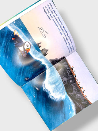An open children's picture book spread showing an illustrated scene of puffins in the ocean with text on the right page, photographed on a light gray background.