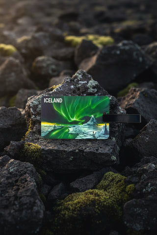 A rectangular luggage tag featuring an illustration of Iceland's northern lights over icy mountains, with a black strap attached on a light gray background.