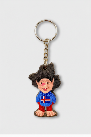 A Troll-themed keychain featuring a small sculpted figure with wild black hair, a pink face, a blue sweater with an Icelandic flag, and red pants, attached to a gold-tone split ring and chain on a light gray background.