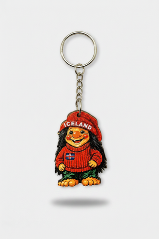 An Iceland-themed cartoon keychain featuring a stylized figure with long black hair, wearing a red knit sweater and matching red hat with 'ICELAND' text, attached to a silver metal key ring and chain on a light gray background.
